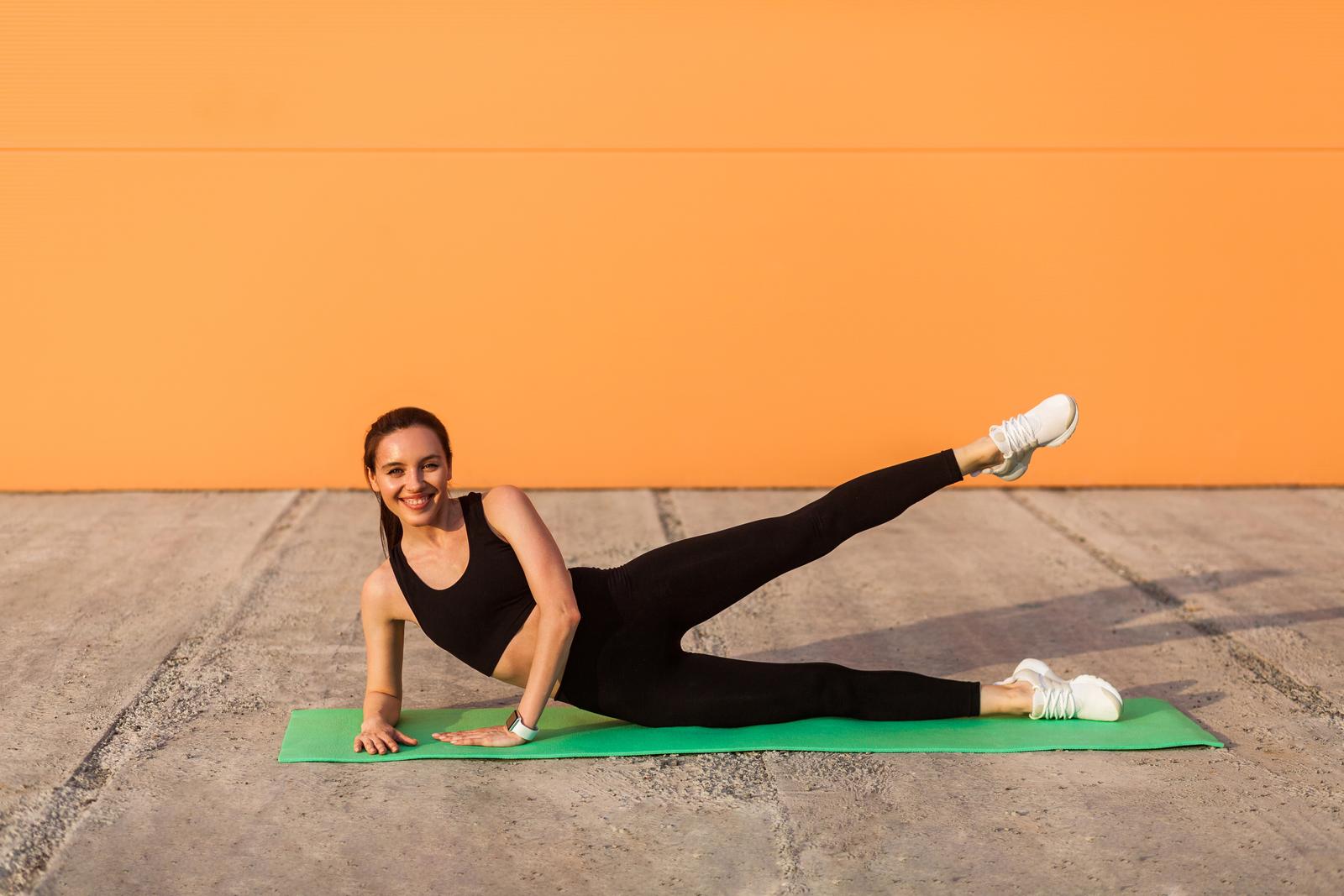 Cheerful athletic girl in tight sportswear, black pants and top, practicing yoga, doing side plank pose with leg lift, stretching muscles, training flexibility. Health care, sport activity outdoor