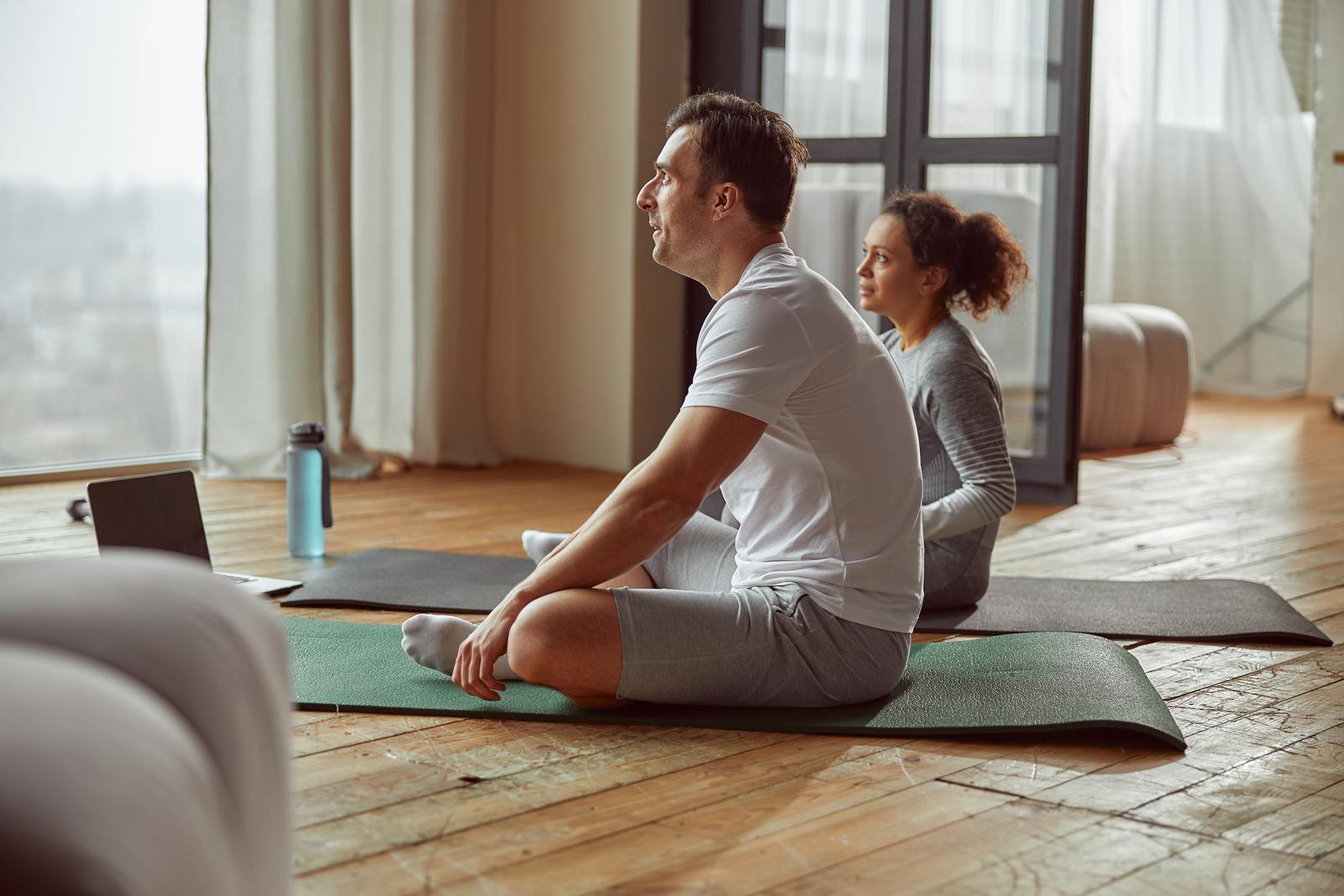 Sporty couple is sitting on mats in front of notebook and doing training together during quarantine. Man and woman doing yoga online at home