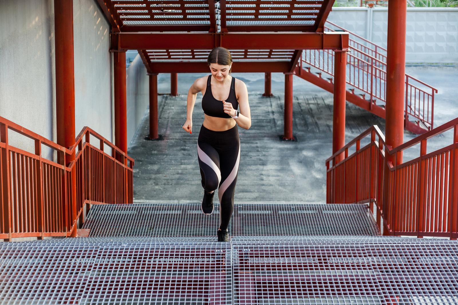 Young brunette woman and sport workout outdoor. Straight view of young athletic slim beautiful woman running up stairs doing cardio interval training in black fashionable sportwear in the street on summer. Outdoor, sport and healthy concept